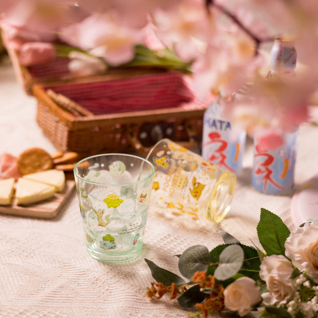 A picnic spread featuring cheese, crackers, flower bouquets, and canned drinks on lace, with Japan Crate’s Doki Doki Kawaii Crate's charming cartoon-printed glasses and pink flowers in the foreground.